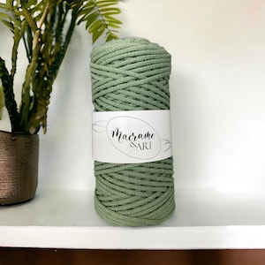 May include: A roll of sage green macrame cord with a white label that reads "Macrame & ART" in a script font. The cord is tightly wound and appears soft. The image is taken in front of a white background.