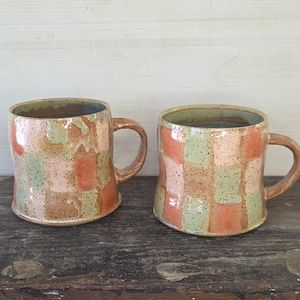 May include: Two handmade ceramic mugs with a patchwork design. The mugs feature a mix of colors, including pink, green, and orange squares. Each mug has a brown handle and a speckled, textured surface. The mugs are displayed on a wooden surface.