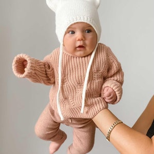 May include: A baby wearing a knitted outfit, including a light pink sweater and matching pants. The outfit is complemented by a white knit hat with cat ears. The baby's outfit is held up by a person's arm.