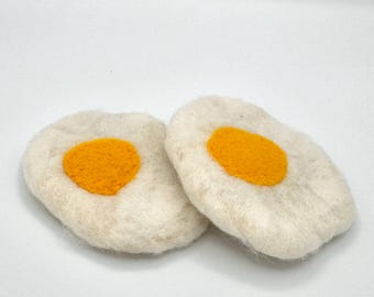 May include: Two felt egg-shaped ornaments with a yellow yolk in the center. The ornaments are white and have a fuzzy texture.