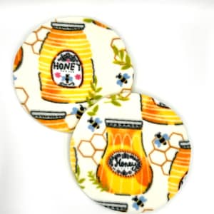 Puede incluir: Dos posavasos redondos de tela blanca con un patrón de tarro de miel y panal. Los tarros son de color amarillo y naranja con la palabra "Honey" impresa en ellos. El patrón de panal es en tonos de amarillo y marrón. También hay pequeñas abejas azules y hojas verdes en los posavasos.