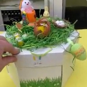 May include: A white gift box with a green grass border and a bunny rabbit design. The box is filled with green grass, a small bunny rabbit, a nest with eggs, and a chocolate egg.