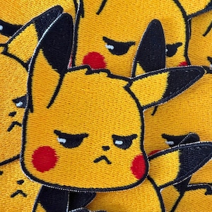 May include: An embroidered patch featuring the yellow face of a cartoon character with long ears tipped in black, narrow eyes, a small nose, and round red cheeks. The character has a grumpy expression with a downturned mouth and black outlines.