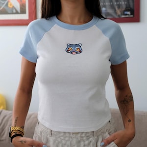 May include: White and light blue raglan t-shirt with a cartoon cat face embroidered on the front. The shirt has a crew neck and short sleeves. The cat face is blue with orange eyes and a pink tongue.