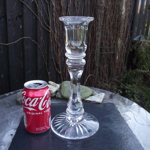 May include: A clear glass candlestick with a tall, slender stem and a wide, fluted base. The candlestick is approximately 25 cm tall and is resting on a dark grey surface.