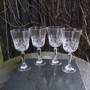 May include: Four clear crystal wine glasses with diamond-cut patterns. The glasses have long stems and round bases, set on a dark grey surface. The background features a dark wooden fence and bare branches.
