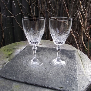 May include: Two clear crystal wine glasses with detailed cut patterns. The glasses have a classic design with a stem and a round base. They are placed on a dark grey slate-like surface, with a natural background of branches.