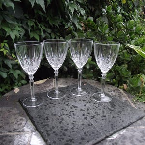May include: Four clear crystal wine glasses with etched patterns, set on a dark grey stone surface. The glasses have long stems and rounded bowls. The background features lush green foliage.