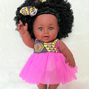 May include: A black baby doll with brown eyes and a pink tulle dress. The doll has curly black hair and is wearing a yellow and black patterned headband.