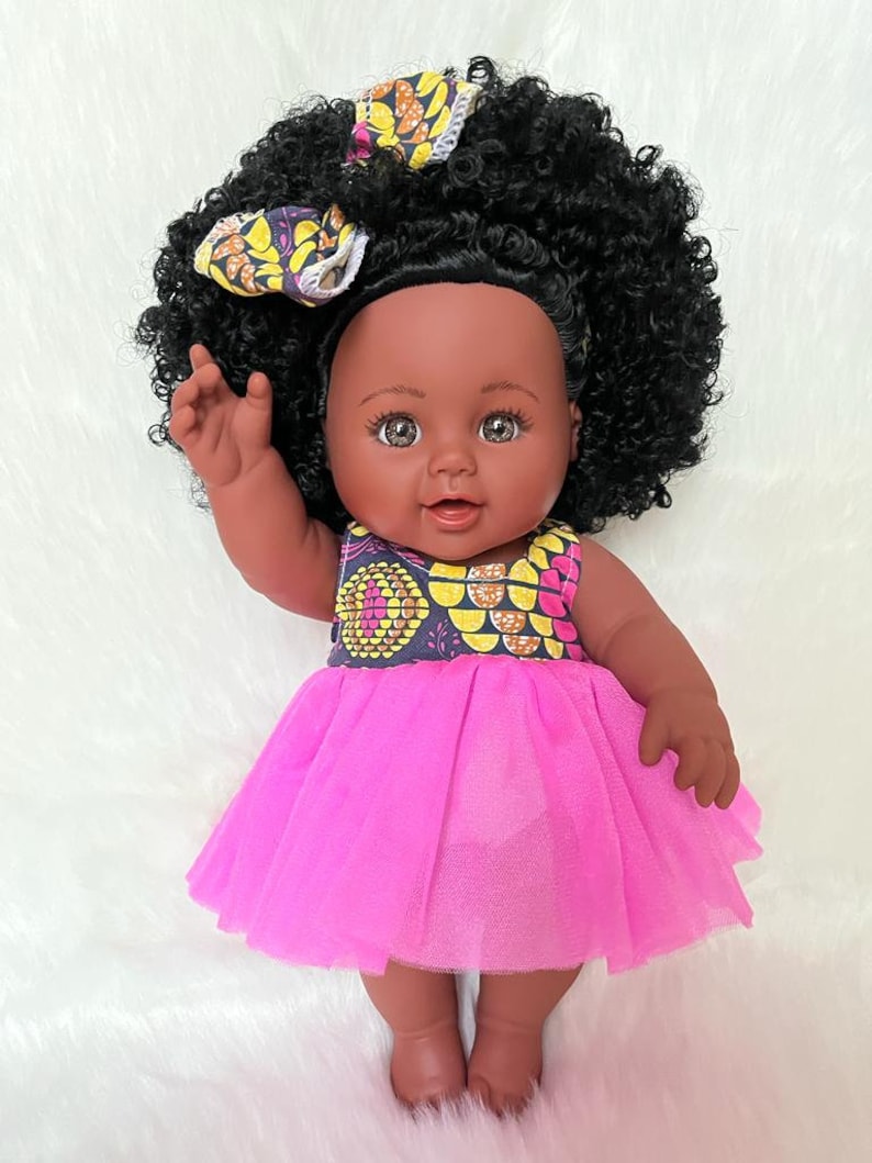 May include: A black baby doll with brown eyes and a pink tulle dress. The doll has curly black hair and is wearing a yellow and black patterned headband.