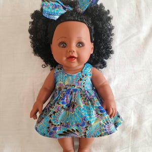 Peut inclure: Poupée bébé à peau foncée avec des cheveux noirs bouclés et un nœud bleu à motifs. La poupée porte une robe à motifs bleus, violets et dorés. La poupée a les yeux bruns et une expression souriante.