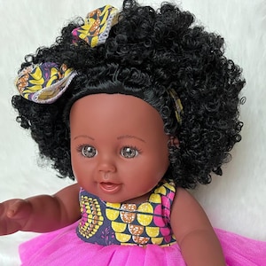 May include: A black doll wearing a pink dress and a yellow and brown patterned headband. The doll has brown eyes and a big smile.