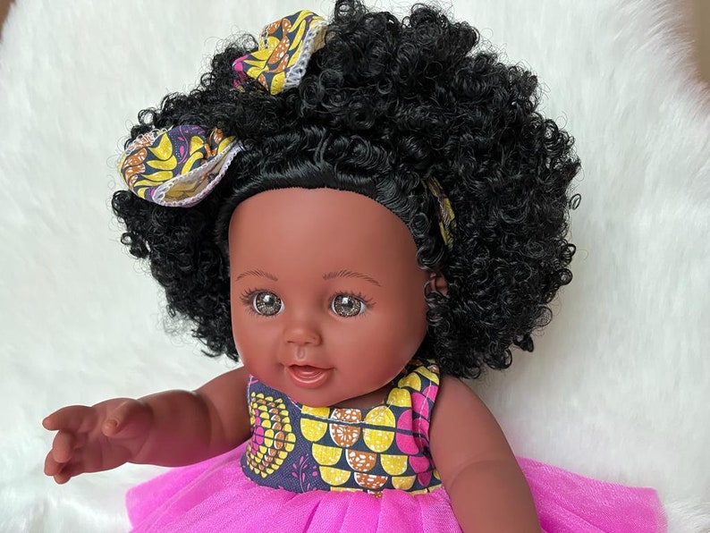 May include: A black doll wearing a pink dress and a yellow and brown patterned headband. The doll has brown eyes and a big smile.