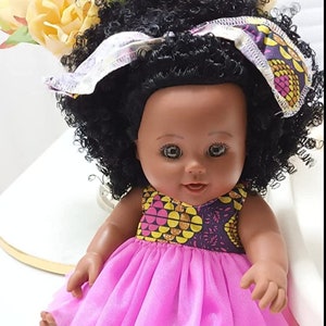 May include: A black doll with brown eyes and a pink tulle skirt. The doll is wearing a yellow, pink and purple patterned dress and a white and purple headband. The doll has curly black hair.