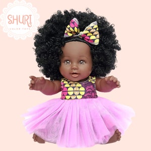 May include: A black doll with brown eyes and a large afro hairstyle. The doll is wearing a pink tulle dress with a yellow and pink patterned bodice. The doll has a pink and yellow patterned bow in her hair.