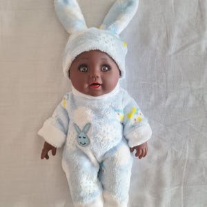 Peut inclure: Poupée pour bébé vêtue d'un chapeau de lapin bleu clair et d'une combinaison assortie. La combinaison a des bordures blanches et un motif de lapin souriant. La poupée a la peau foncée et les yeux bruns.
