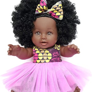 May include: A black baby doll with brown eyes and a pink tulle dress. The doll has a large afro wig and a pink and yellow patterned headband with a bow.
