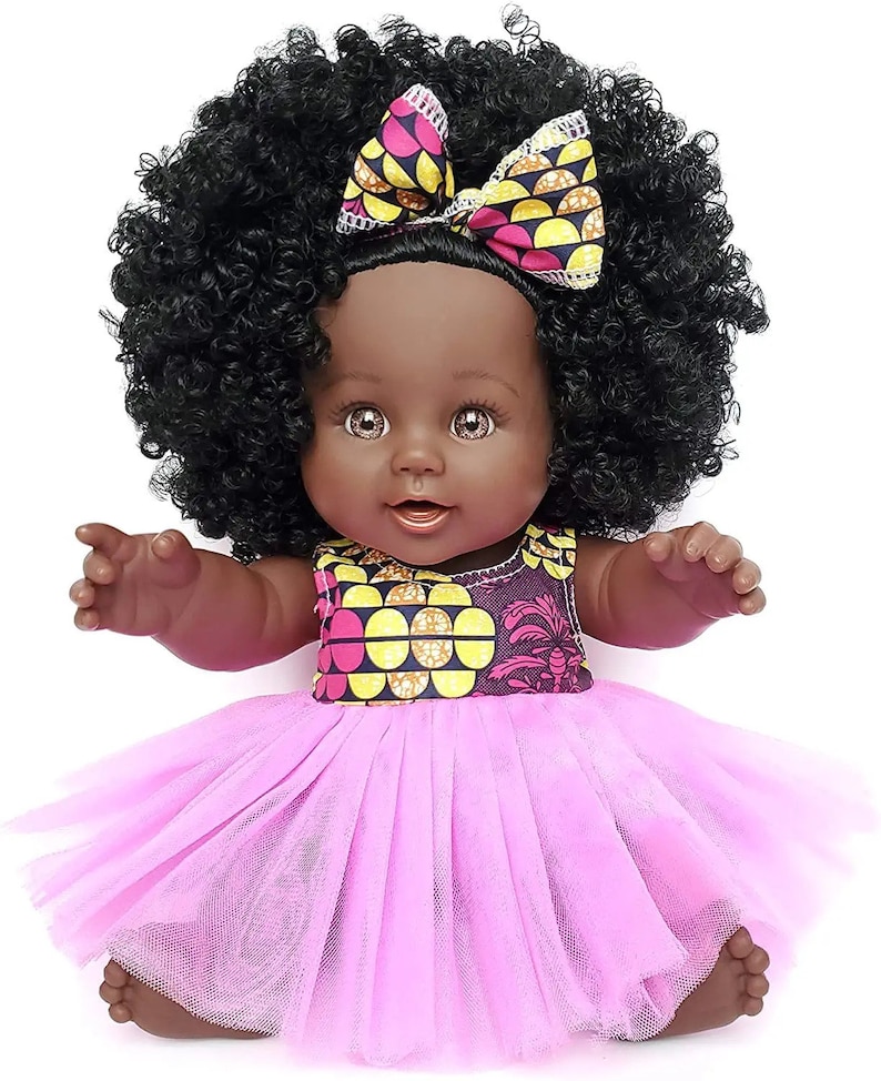 May include: A black baby doll with brown eyes and a pink tulle dress. The doll has a large afro wig and a pink and yellow patterned headband with a bow.