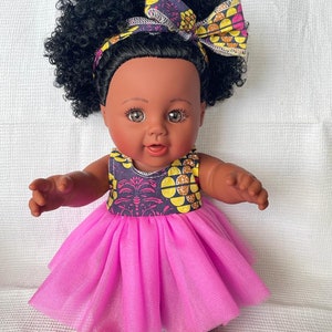 May include: A black doll with brown eyes and a pink tulle dress. The doll has a large afro hairstyle and is wearing a headband with a yellow and purple floral pattern.