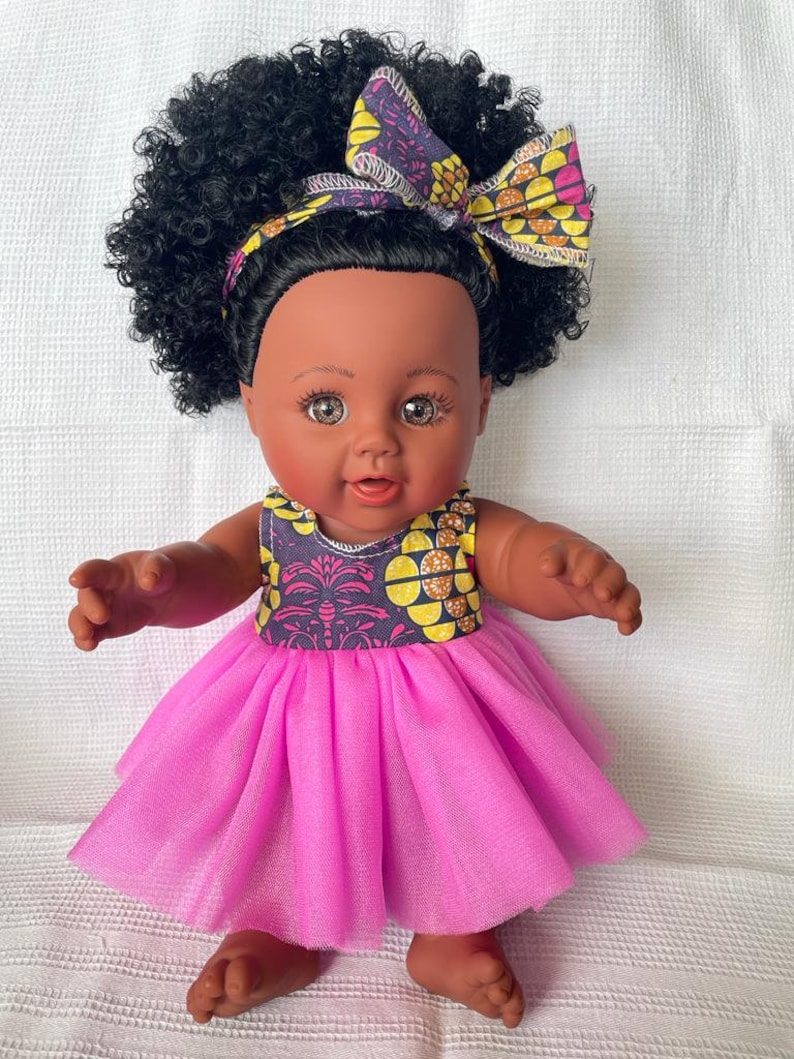 May include: A black doll with brown eyes and a pink tulle dress. The doll has a large afro hairstyle and is wearing a headband with a yellow and purple floral pattern.