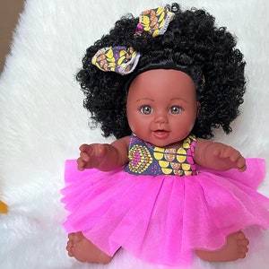 May include: A black doll with brown eyes and a pink tulle dress. The doll has a yellow and brown patterned headband and curly black hair.
