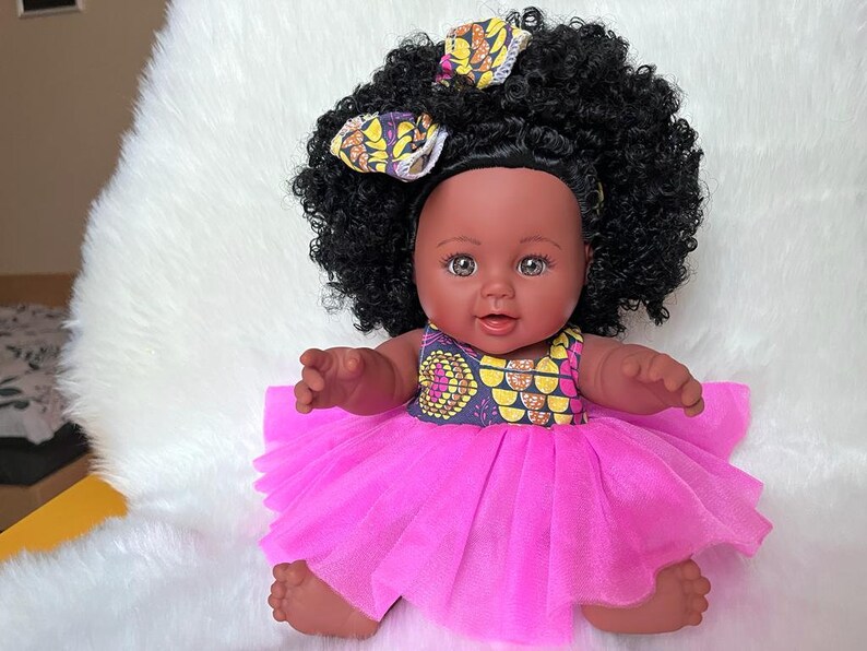 May include: A black doll with brown eyes and a pink tulle dress. The doll has a yellow and brown patterned headband and curly black hair.
