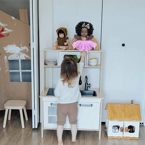May include: A child is playing with a wooden toy kitchen set. The kitchen has a sink, oven and shelves. There are two dolls on the top shelf, one with brown hair and a brown dress and the other with black hair and a pink dress. The child is wearing a white shirt and brown trousers.