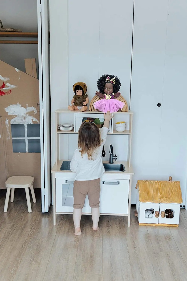 May include: A child is playing with a wooden toy kitchen set. The kitchen has a sink, oven and shelves. There are two dolls on the top shelf, one with brown hair and a brown dress and the other with black hair and a pink dress. The child is wearing a white shirt and brown trousers.