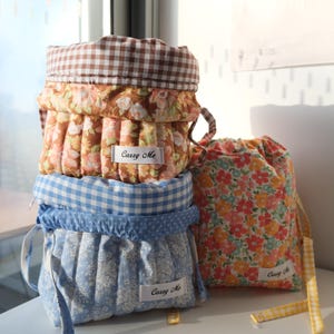 May include: Three fabric drawstring pouches with quilted textures. Two are stacked, featuring brown gingham, floral patterns, and blue gingham with floral prints. A third pouch stands beside them with a vibrant floral print and yellow gingham ties.