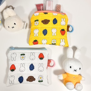 May include: Several small, quilted pouches and soft toys are shown. A yellow pouch is patterned with cartoon bunnies, flowers, and fruit. A white pouch has a similar design. A white bunny keyring and a plush bunny in an orange outfit are also present.