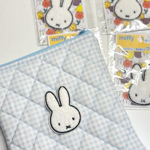 May include: A light blue and white gingham quilted pouch featuring a white Miffy rabbit patch with black outlines. Three additional Miffy rabbit patches in individual clear plastic packaging are visible in the background, each displaying the text miffy.