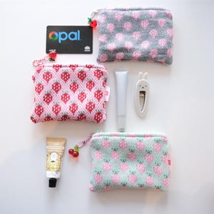 May include: Three rectangular knit pouches featuring strawberry patterns in grey, pink, and mint green. Each pouch has a cherry-shaped zipper pull. Included items are an Opal transit card, a silver tube, a white rabbit hair clip, and a small cream tube.