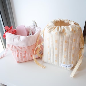 May include: Two quilted drawstring pouches with a floral pattern and a white label reading Carry Me. One pouch is light pink with a pink gingham interior, and the other is cream with a yellow gingham drawstring and interior lining.