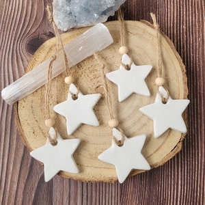 May include: Six white ceramic star ornaments with twine and wooden beads hanging from a wooden slice. The ornaments are arranged in a circle on the wooden slice.