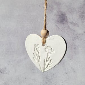 May include: A white heart-shaped decoration featuring embossed floral patterns, including a dandelion and other plants. It is suspended by a natural twine string with a wooden bead. The background is a soft, mottled grey.