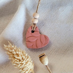 May include: A pink heart-shaped ceramic ornament with a leaf pattern hanging from a twine string with wooden beads.