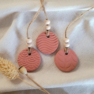 May include: Three terracotta clay ornaments with different embossed patterns, each hanging from a twine cord with wooden beads. The ornaments are a light reddish-brown colour.
