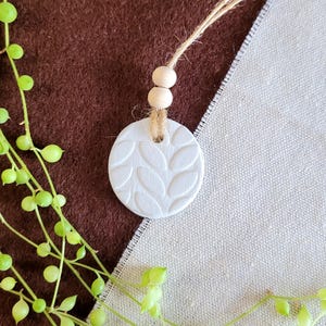 May include: A white, circular essential oil diffuser with a leaf pattern, suspended by a natural twine cord and wooden beads. The diffuser is set against a brown and beige fabric background, with a green plant in the foreground.