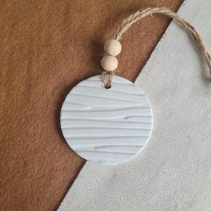 May include: White ceramic ornament with a woodgrain texture, hanging from a twine string with two wooden beads.