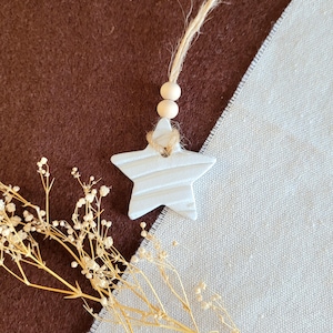 May include: A white ceramic star ornament with a textured surface and a natural twine hanger. The star is hanging from two wooden beads on a twine string.