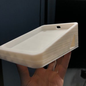 May include: White 3D printed phone stand with a slot for charging cables. The stand has a slightly angled surface to prop up a phone.