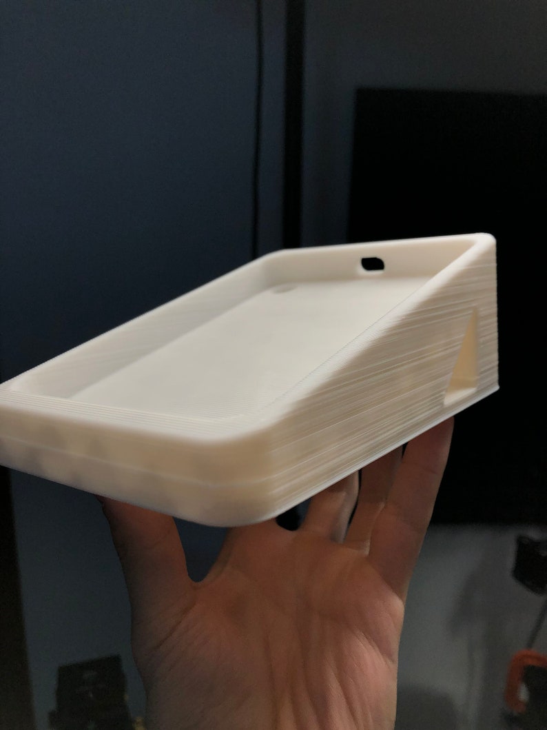 May include: White 3D printed phone stand with a slot for charging cables. The stand has a slightly angled surface to prop up a phone.