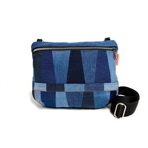 Könnte beinhalten: Eine rechteckige Umhängetasche aus Patchwork-Denim in verschiedenen Blautönen. Die Tasche verfügt über einen Reißverschluss an der Oberseite, eine horizontale Naht auf der Vorderseite und einen verstellbaren schwarzen Gurt mit Metallschnalle.