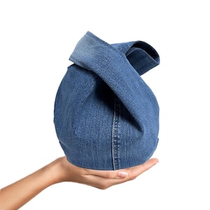 Könnte beinhalten: Eine Jeans-Handtasche mit einem einzigartigen Knotendesign. Die Tasche besteht aus blauem Jeansstoff und hat eine runde Form. Der Griff wird durch Verdrehen und Verknoten des Jeansmaterials gebildet. Die Tasche wird in der Hand einer Person gehalten.