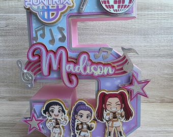 May include: A multi-layered, decorative number five with a music theme. Features the name "Madison", musical notes, stars, and cartoon figures. Includes the text "HUNTRIX" and a disco ball design. Predominantly pink, blue, and silver.