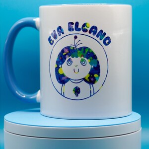 May include: White ceramic mug with a blue handle featuring a cartoon illustration of a girl with blue and yellow hair and the text "EVA ELCANO" in blue. The mug is on a light blue surface.