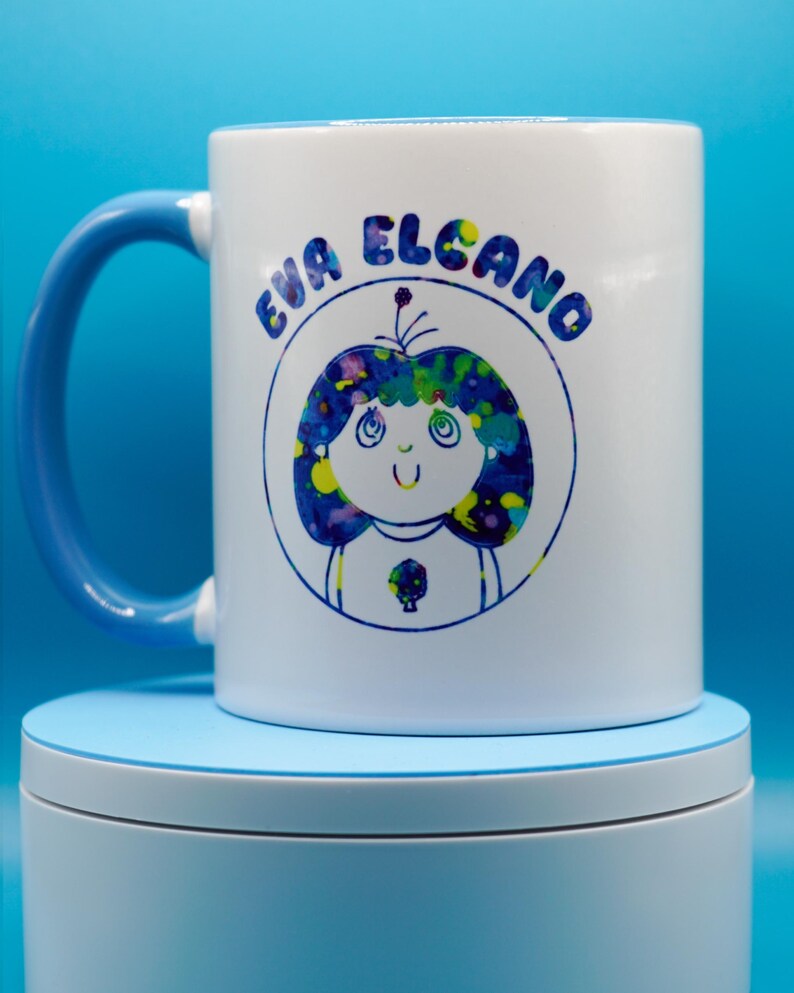 May include: White ceramic mug with a blue handle featuring a cartoon illustration of a girl with blue and yellow hair and the text "EVA ELCANO" in blue. The mug is on a light blue surface.