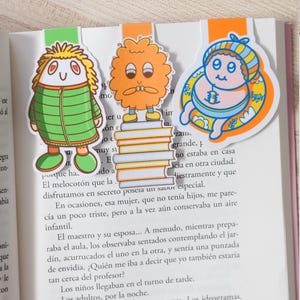 May include: Three colorful cartoon character bookmarks. The bookmarks feature a green figure, an orange figure, and a blue baby-like figure. Each bookmark is inserted into the pages of a book.