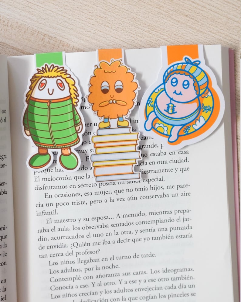 May include: Three colorful cartoon character bookmarks. The bookmarks feature a green figure, an orange figure, and a blue baby-like figure. Each bookmark is inserted into the pages of a book.