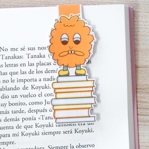 Op de afbeelding: Oranje bladwijzer met een stripfiguur op een stapel boeken, ingevoegd in een boek. Het personage heeft een bezorgde uitdrukking, een rond lichaam, twee ogen en kleine armen. Het boek heeft Spaanse tekst.